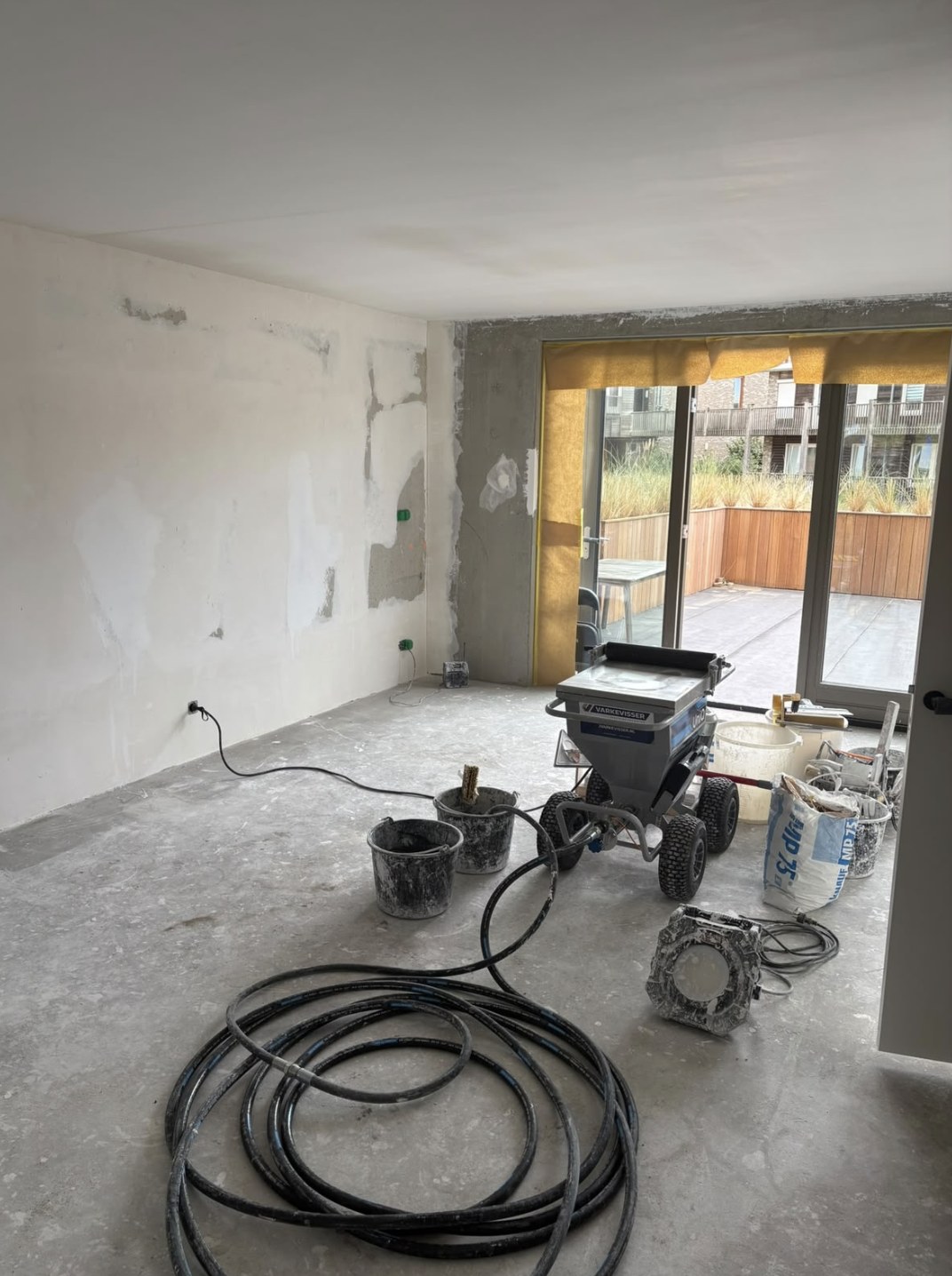 Nieuwbouwkamer tijdens het werk, met de dunne laag op de wand en de pleistermachine op de vloer